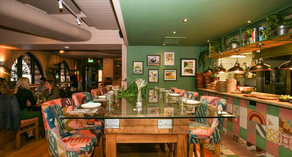 A long wooden table with a glass top and patterned upholstered chairs beside the tiled open kitchen at The Granary