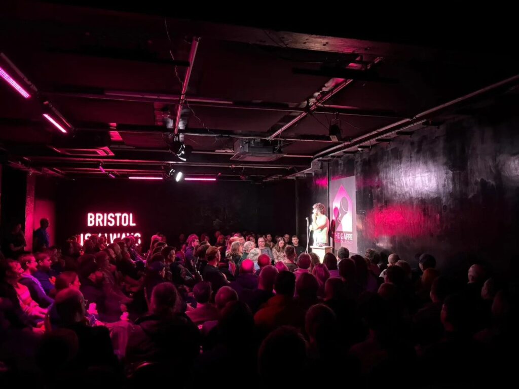 A packed audience watching a comedian on stage under pink lights at The Gaffe in Bristol